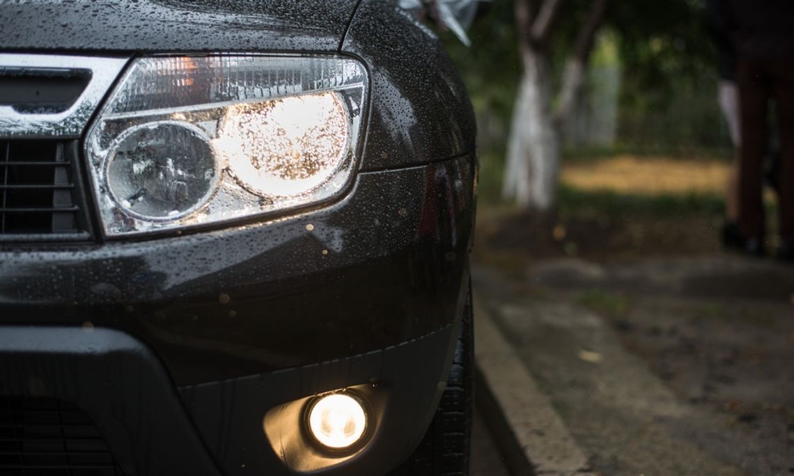 A black car with rain drops on its hood and bumper, showing an illuminated headlight and fog light outdoors.