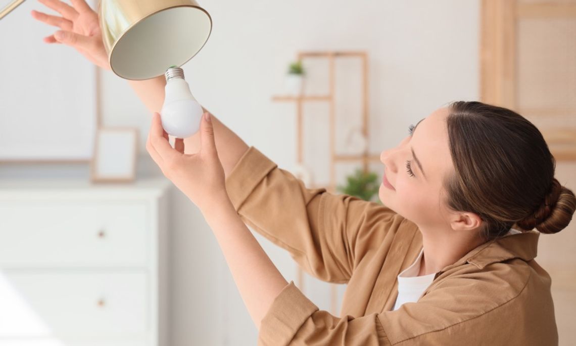 Common Causes of Flickering Household Lights A woman in a brown shirt replacing a light bulb in a gold lamp, holding the fixture with one hand in a bright, modern home.
