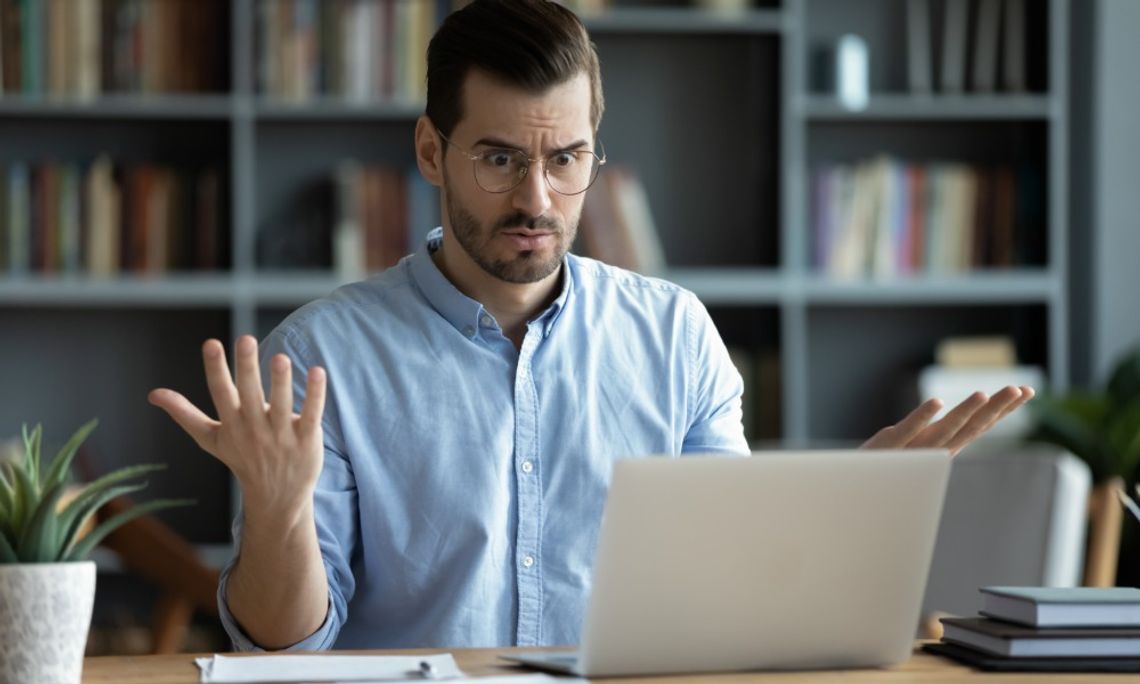 Common Computer Parts Offices Replace Most Often A man wearing a blue button-down shirt is raising his hands in frustration while looking at his computer.