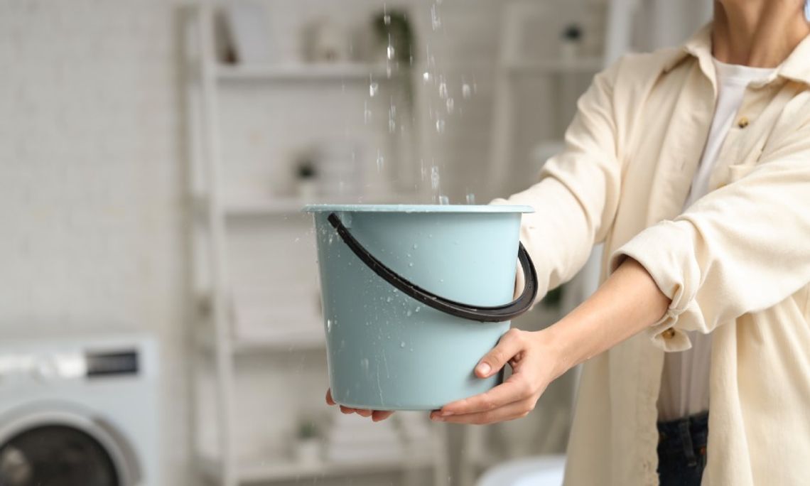 In a laundry room, a woman is holding a blue bucket to catch drops of water falling from a leak in the ceiling.