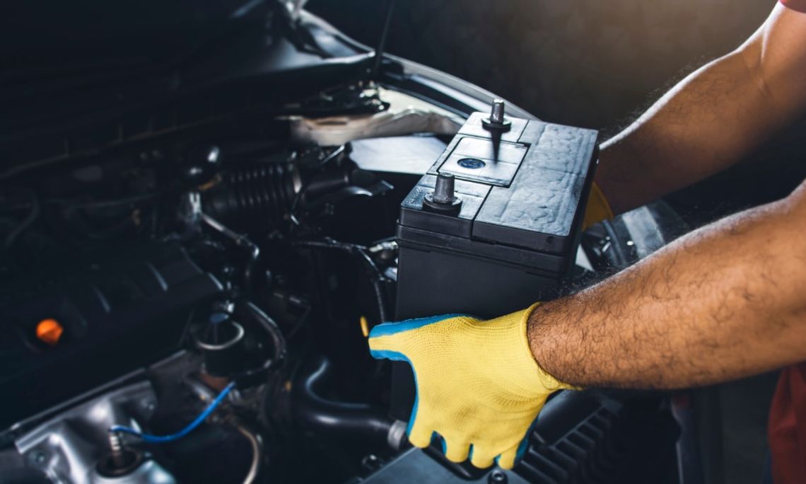 Common Maintenance Mistakes With Older Cars A close-up of a person holding a car battery inside an open engine compartment with tools and cables nearby.