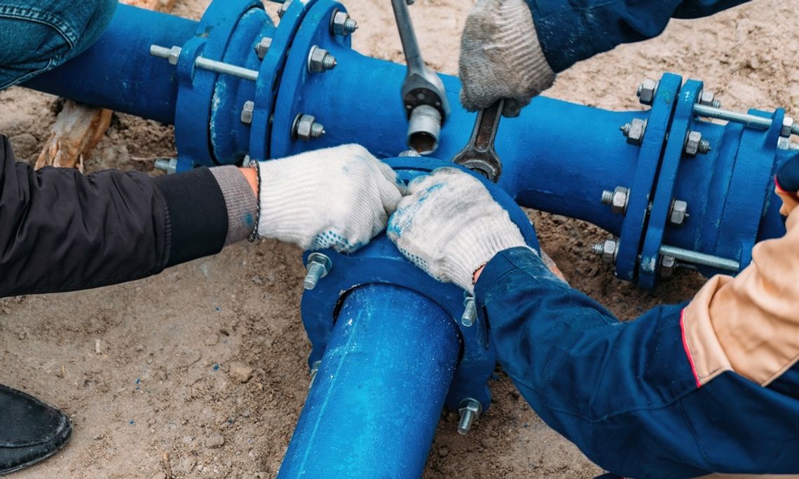 Two workers wearing gloves, loosening the bolts of a blue pipeline. One has a regular wrench and the other has a socket wrench.