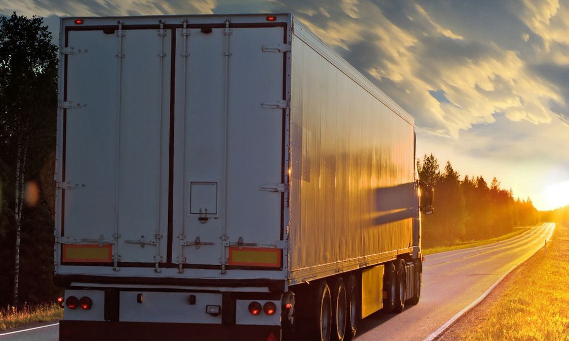 A semi-truck driving down a one-lane rural highway with trees nearby. The sun is rising at the end of the horizon.