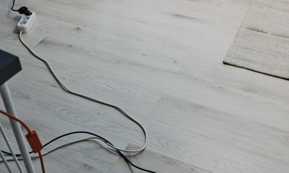 Common Tripping Hazards To Prevent at Home A tan rug on a wooden floor inside a home. A long extension cord runs across the floor near the rug.