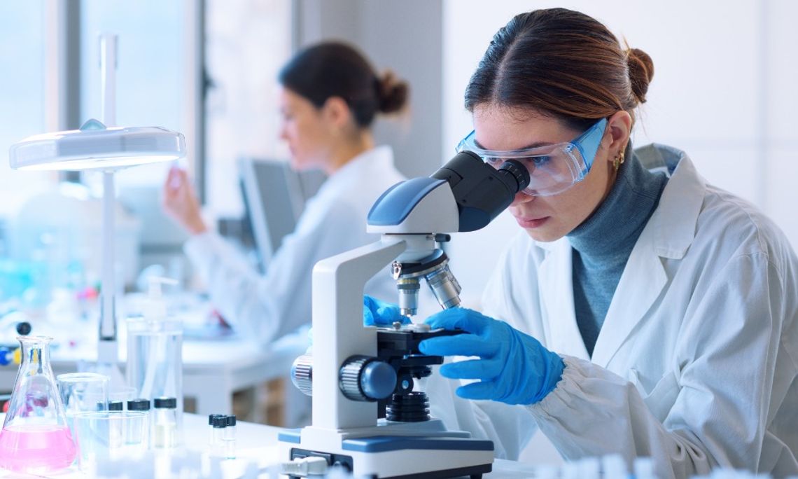 Connecting Particle Properties to Product Performance A lab scientist wears a white coat, blue latex gloves, and goggles as they work with a microscope to review particles.