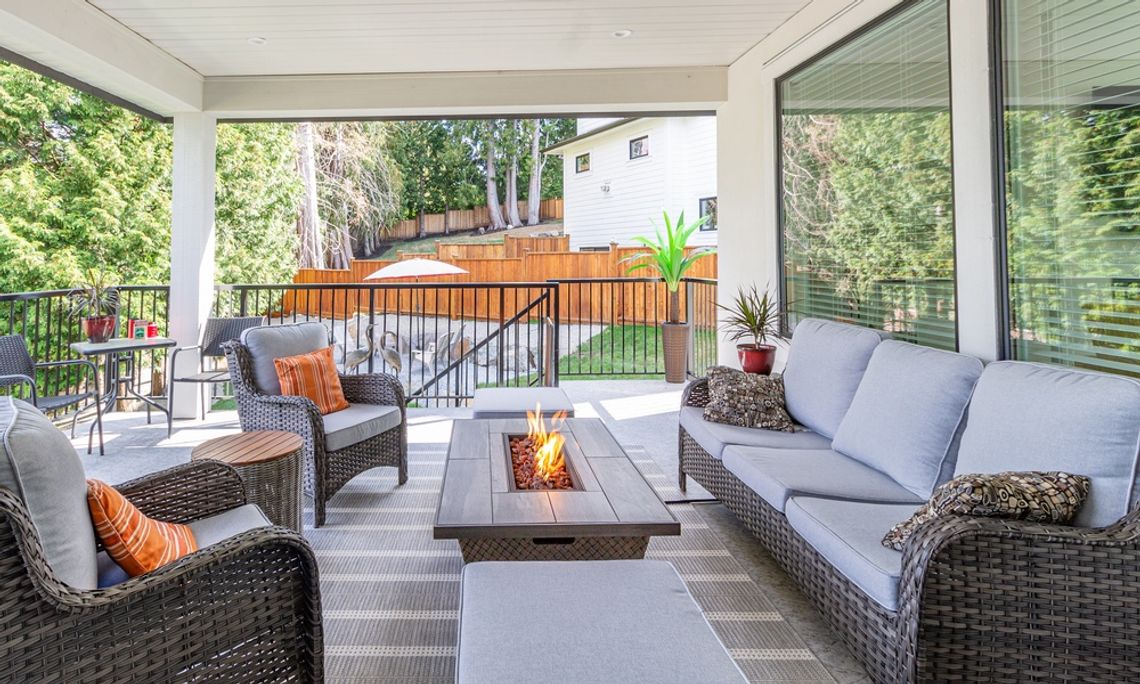 A covered patio has matching brown wicker furniture with blue-gray cushions. A fire burns in the rectangular fire pit table.