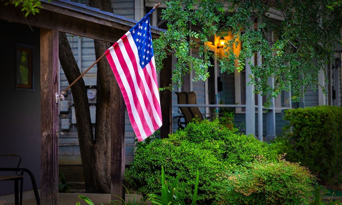 An American flag hangs off the end of a flagpole attached to the pillar of a wooden house. Greenery surrounds the house.