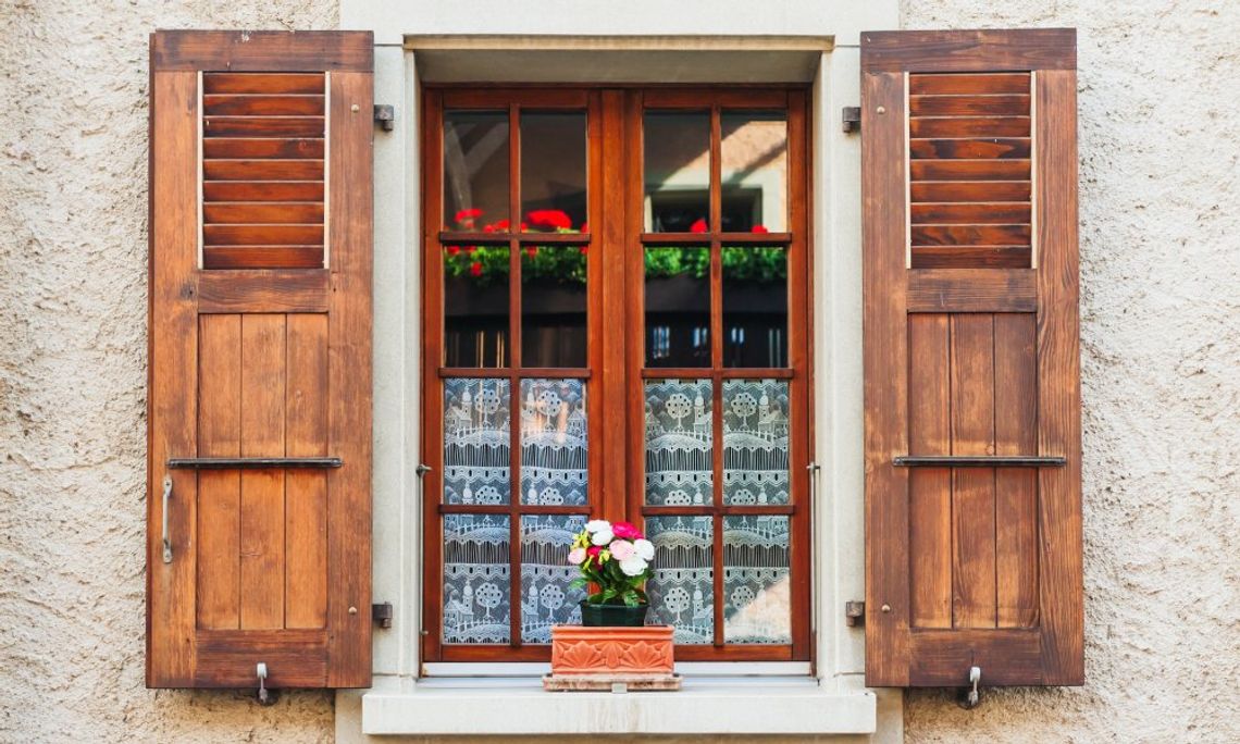 A home’s exterior window, up close. It has custom wood shutters, white lace curtains, and a small window box with flowers.