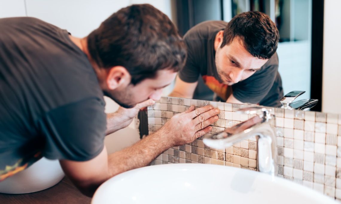 DIY Renovations for a Functional and Mold-Free Bathroom A man placing the backsplash of a sink inside a bathroom with a large mirror showing the reflection of the man.