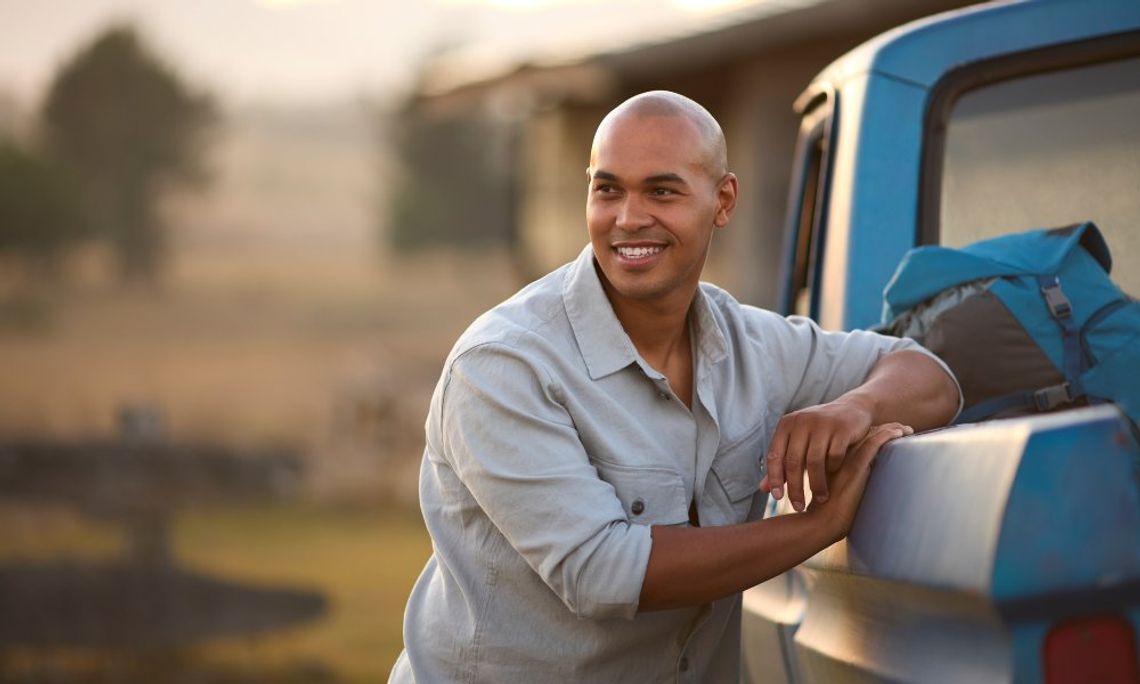 A smiling bald man leans against the left side of his blue pickup truck outside. A backpack is in the truck bed.