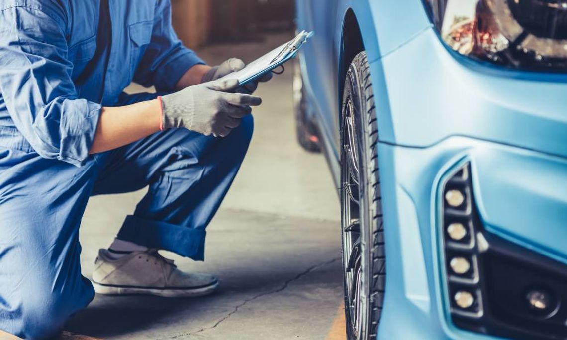 Early Warning Signs That Your Vehicle Needs Maintenance A mechanic in a blue jumpsuit kneeling next to the wheel of a blue car. He is holding a clipboard in his hand.