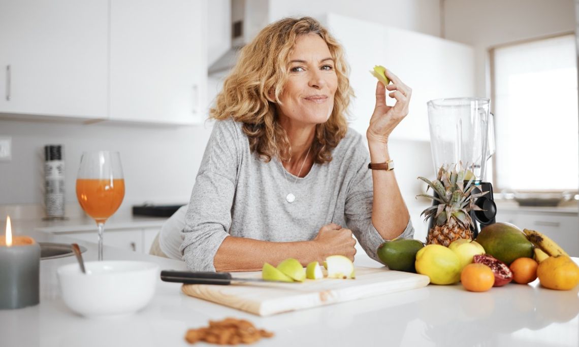 A person eating a slice of fruit in their kitchen. An assortment of fruit is on the counter in front of them.