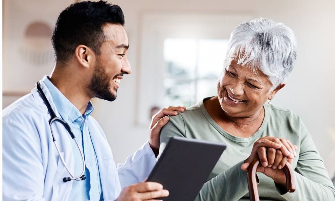 A young male doctor shows a senior female patient something on his tablet. They are smiling and happy.