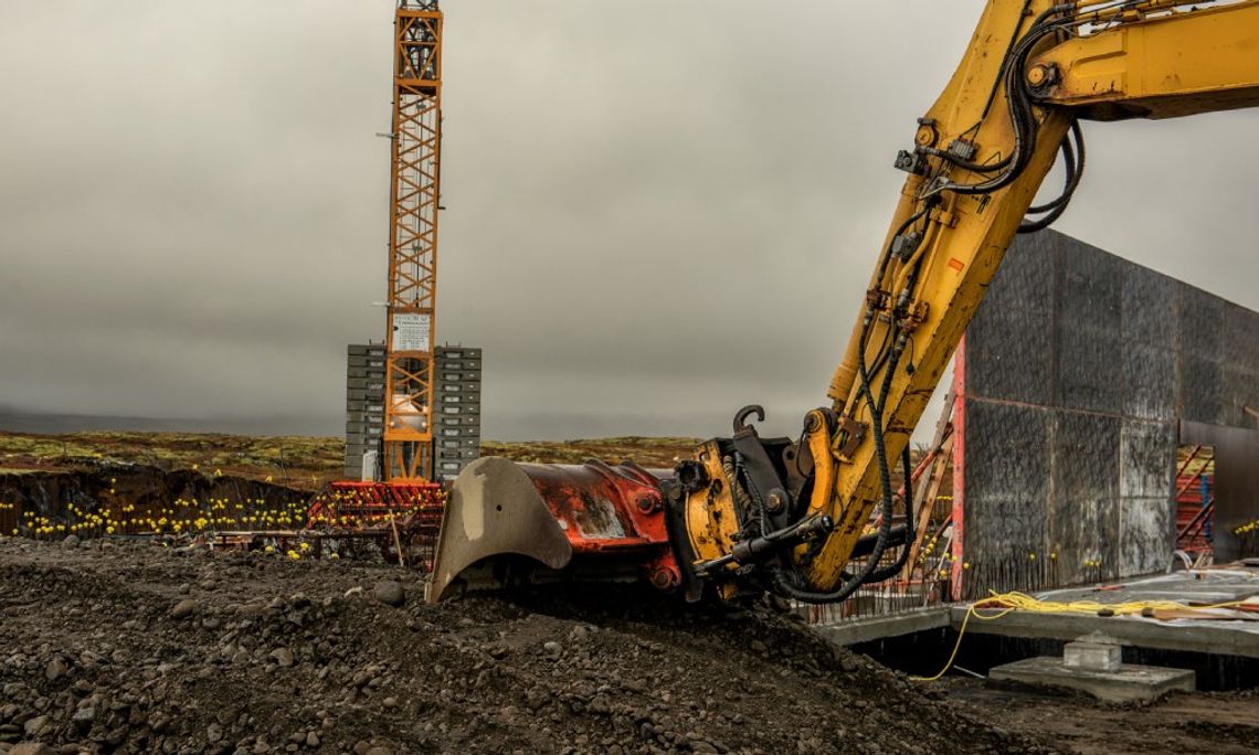 A yellow excavator lifts dark soil at a construction site, with a crane and unfinished building in the background.
