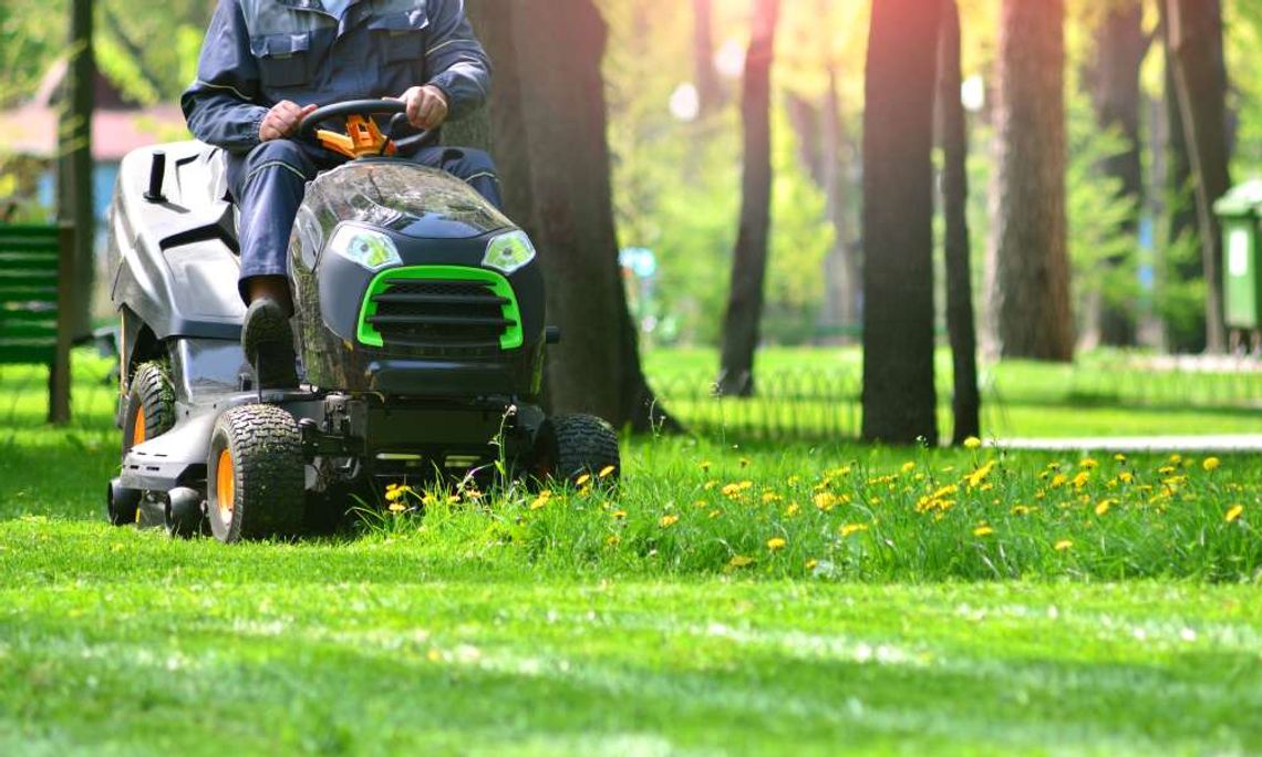 A man in a blue jumpsuit riding on a lawnmower. He is currently driving through a patch of tall grass and yellow flowers.