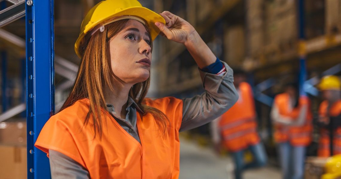 A woman wearing a yellow hard hat and an orange safety vest is standing in a warehouse. She has a hand on the brim of the hat.