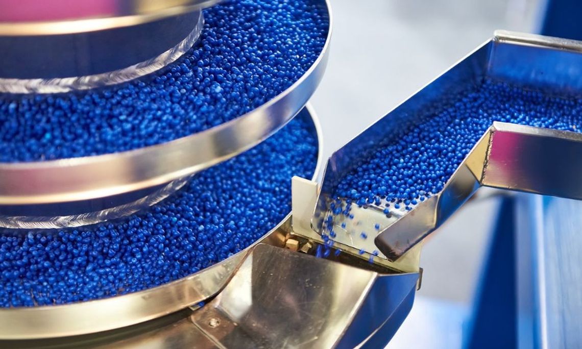 A close-up view shows thousands of tiny blue plastic pellets traveling along a stacked metal track.