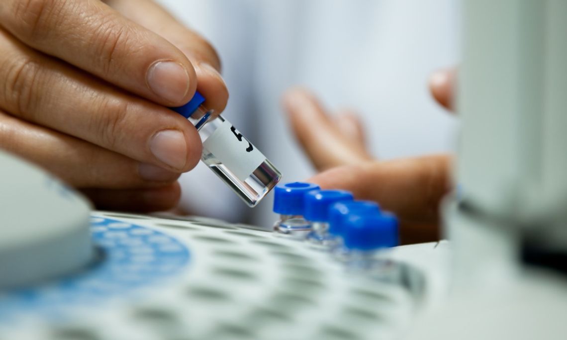 A close-up of a scientist's hand lifting a small labeled glass vial out of a chromatograph in a lab.