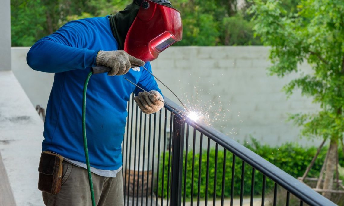 Gathering the Right Gear for Your Home Welding Project A man wearing a welding helmet and gloves working on welding the steel frame of a black fence outdoors.