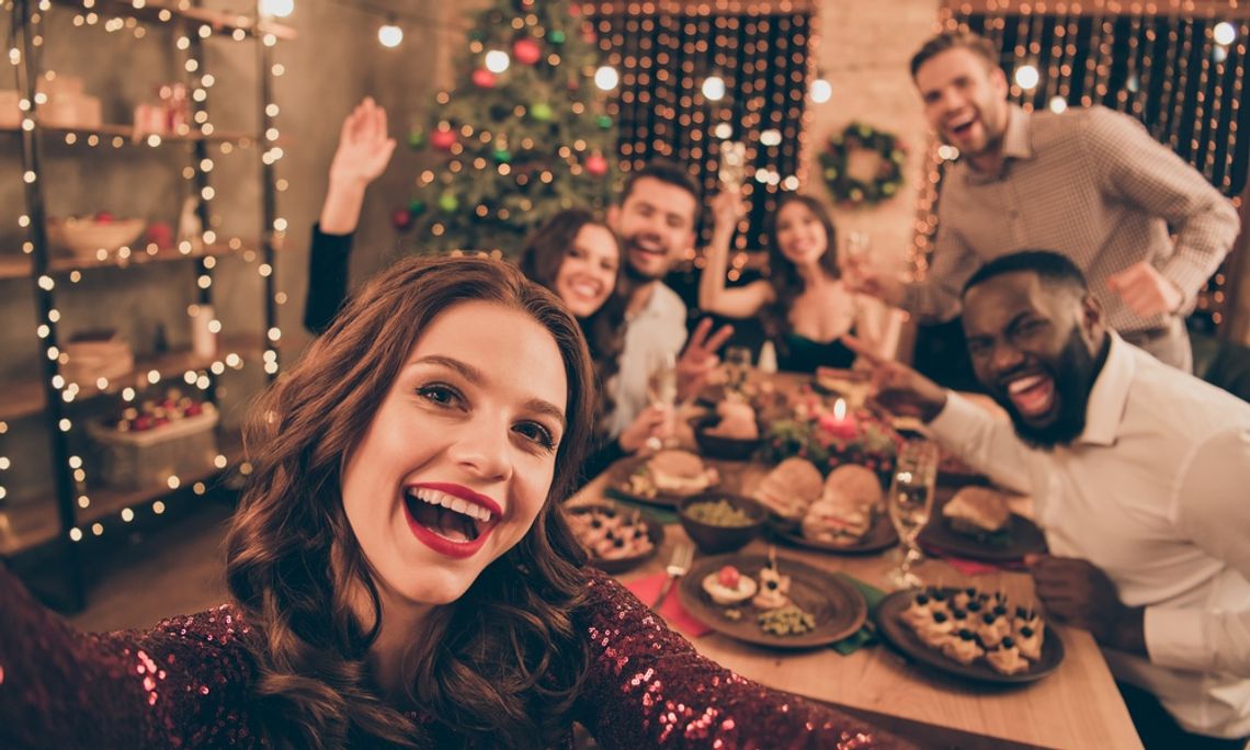 A woman takes a selfie with her party guests in the photo behind her. They're gathered for a holiday party around the table.
