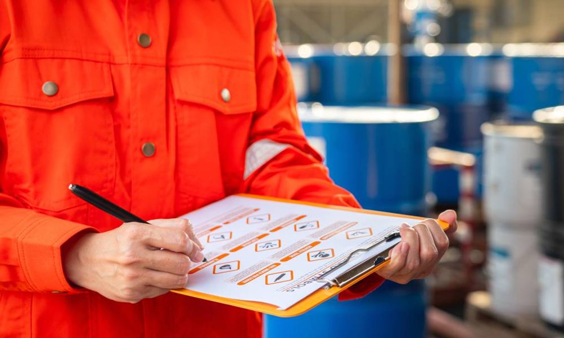 A safety officer in a bright orange jumpsuit holds and consults a checklist. Several blue barrels are in the background.