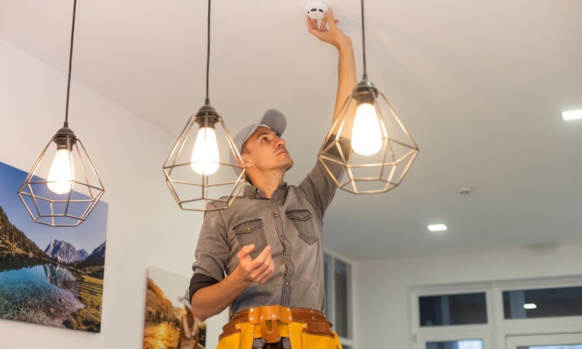 A guy wearing a gray hat and a yellow tool belt installs a new smoke detector on the ceiling of a home.