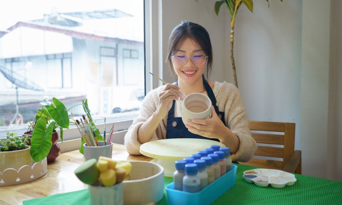 Hobbies To Take Up That Are Great for Mindfulness A woman sitting at a table using a paintbrush to paint on the surface of a clay mug. Other art supplies are on the table.