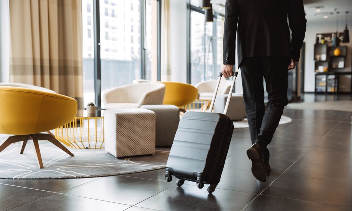 Business traveler pulls a rolling suitcase through a modern hotel lobby with yellow chairs and large windows.