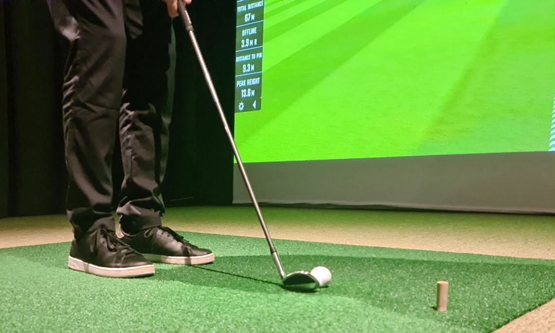 How an Indoor Golf Simulator Can Be Good for Your Health A ground-level close-up of a man's feet behind a golf club wedge and a ball on a tee box inside a golf simulator.