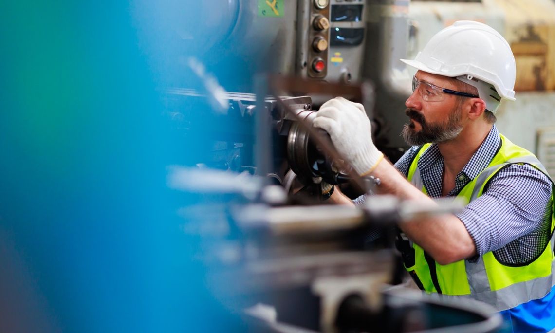 A man is wearing safety goggles, white gloves, and a hard hat as he's kneeling beside and working on a metal lathe machine.