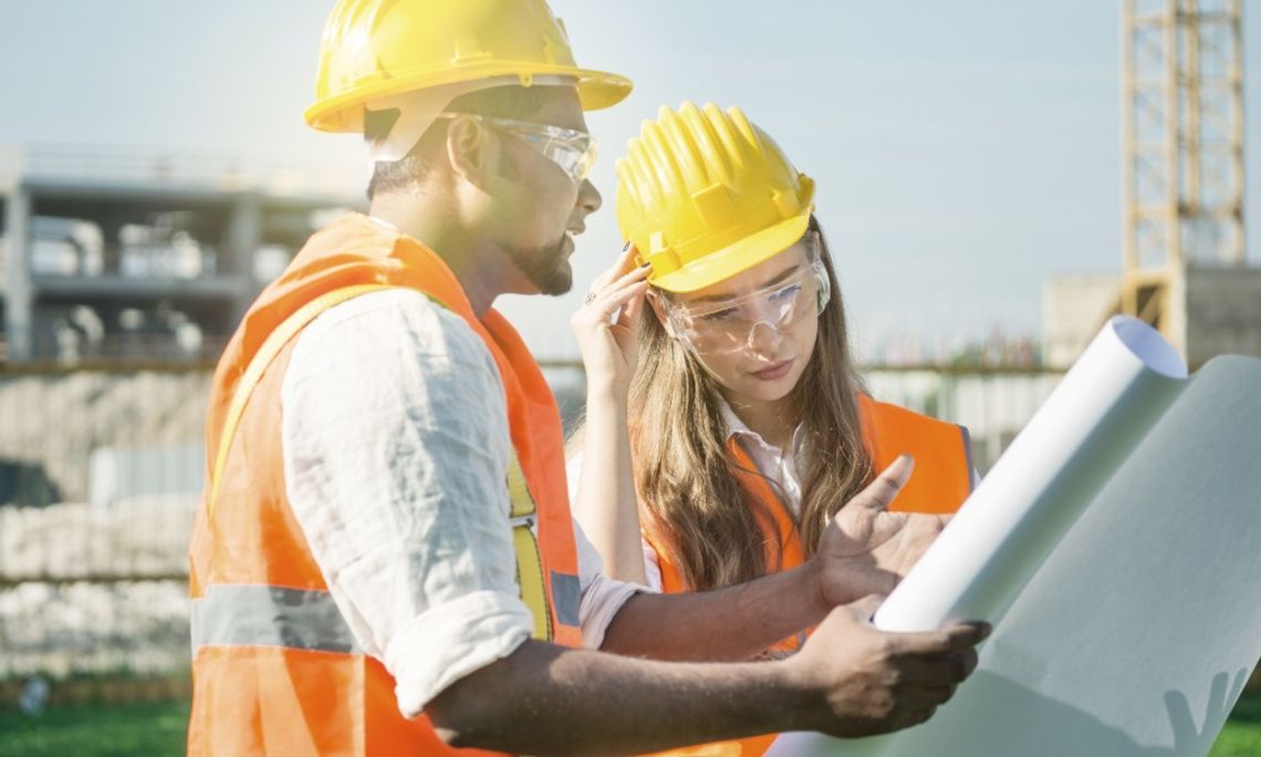 How Changing Project Phases Affect Safety Staffing A man and a woman wearing high-visibility safety vests, hard hats, and safety goggles looking at documents outside.