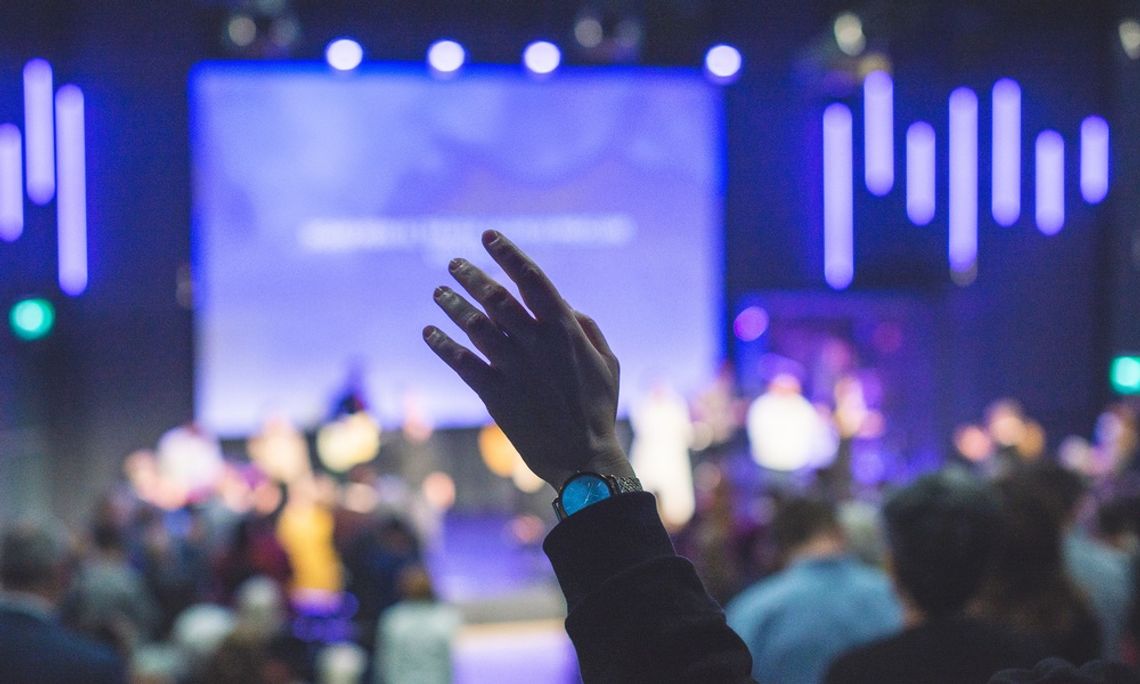 A close-up of someone's hand raised during church service. The stage lights are bright and blue as they worship.