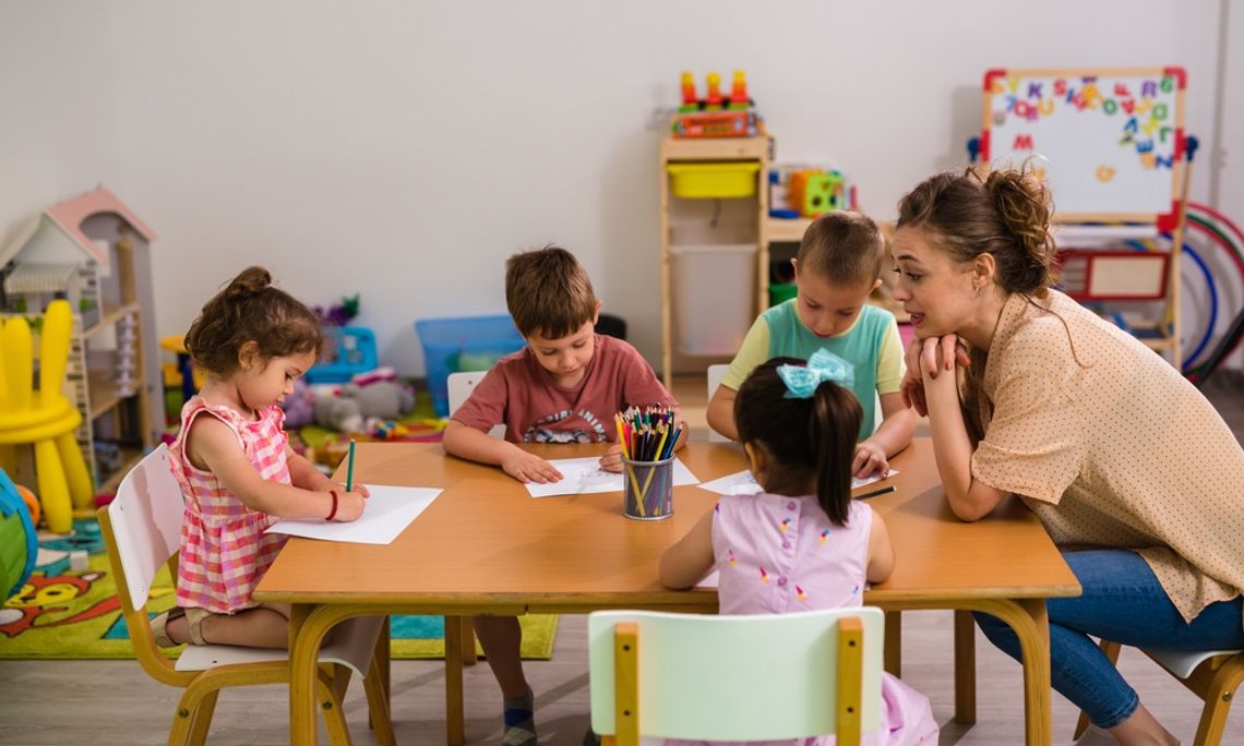 How Daycare Staff Can Encourage Group Play A daycare teacher guides five young children drawing at a classroom table with colored pencils and educational toys.