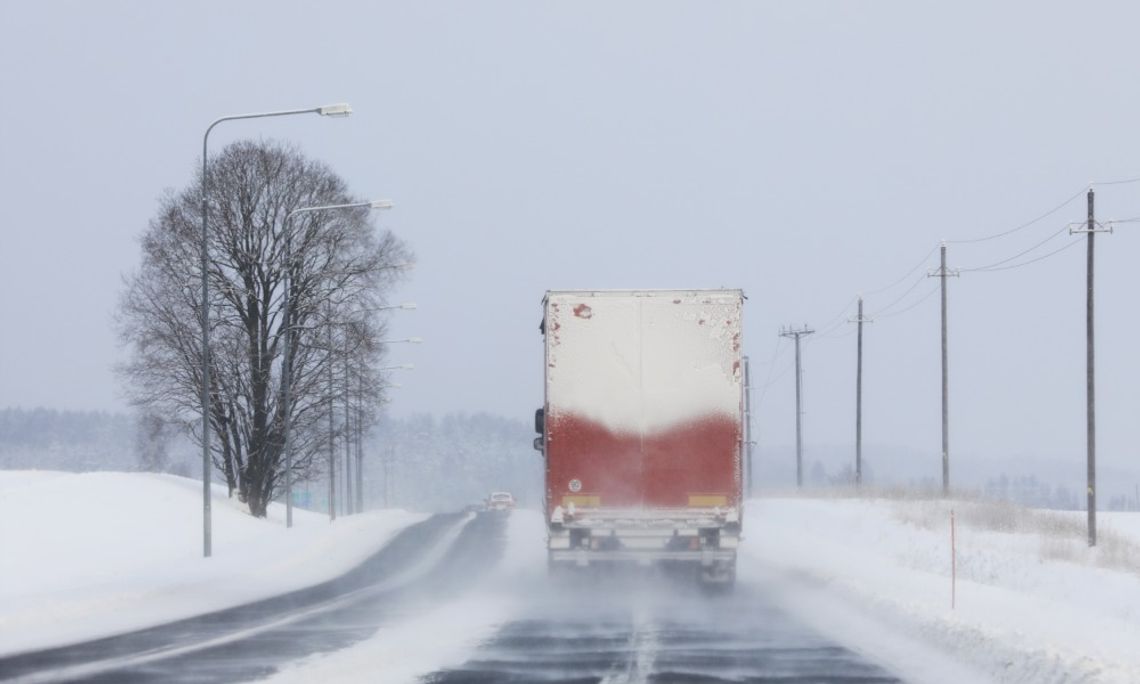 A semi truck driving on a snow-covered highway during snowfall, with trees, streetlights, and power lines along the road.