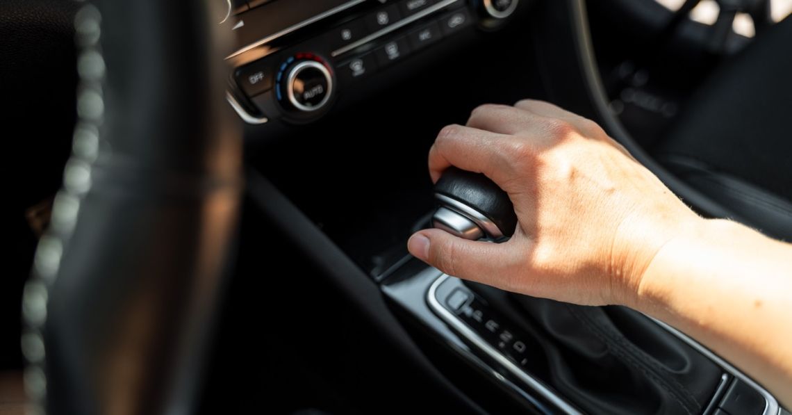 How Does Gear Shifting in a Car Actually Work? A hand holding a black gear shift in a car, with dashboard controls and sunlit center console details visible.