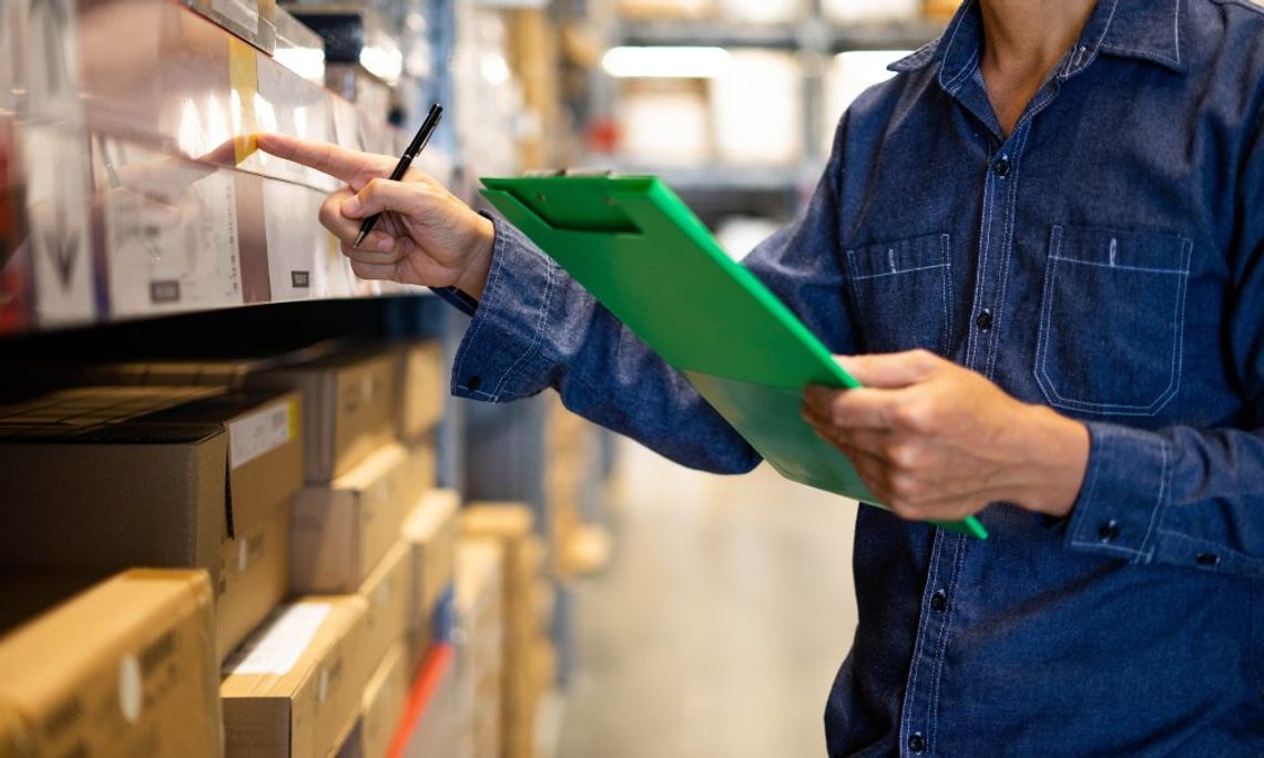 A person in a denim shirt holding a green clipboard and examining various boxes that are stacked on warehouse shelves.