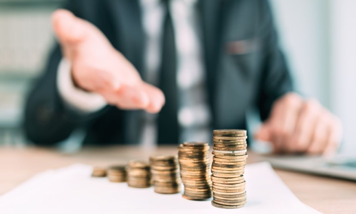 A blurred man in a suit sitting behind a desk and gesturing with one hand to a row of increasingly tall coin stacks.