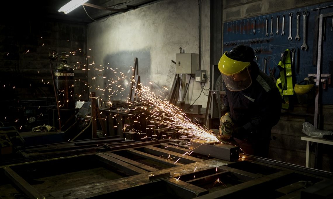 A worker wearing a face shield and gloves grinding a metal frame as bright sparks scatter across a dark workshop.