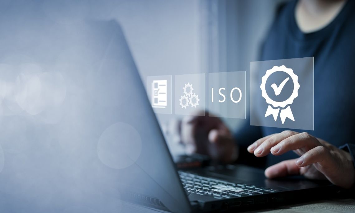 A close-up of a woman using a laptop with holographic icons for a checkmark badge and "ISO" floating in front of her.