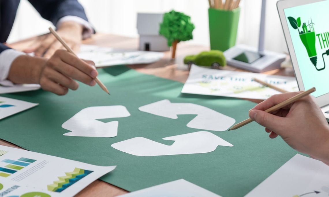 Hands sketch recycling icon during eco planning meeting, with charts, reports, and laptop displaying “Think Green” message.