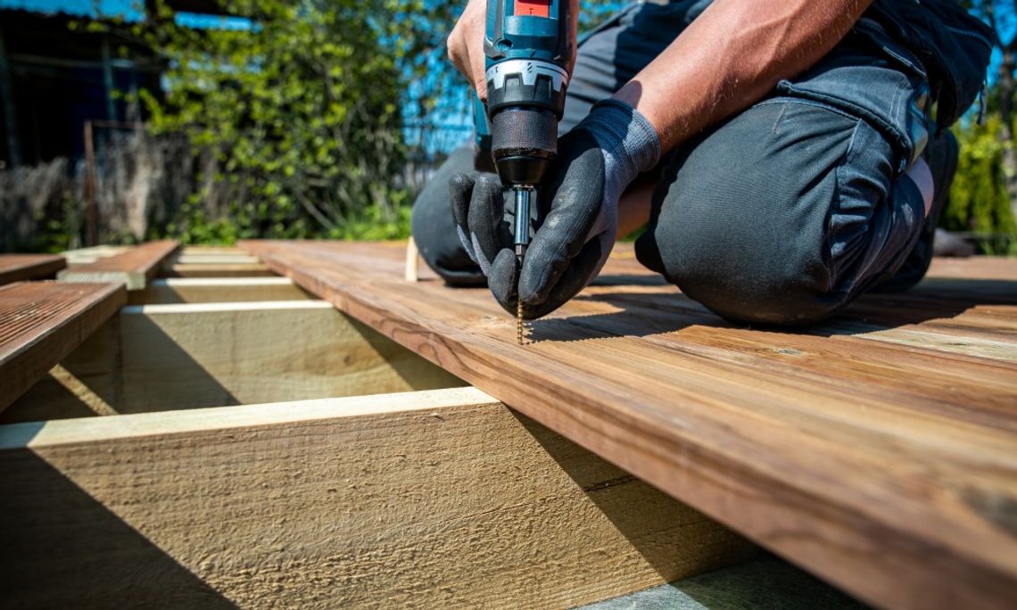 How Long Does a Wooden Deck Typically Last? A close-up of a person using an electric drill to fasten a screw into wooden deck boards over exposed framing.