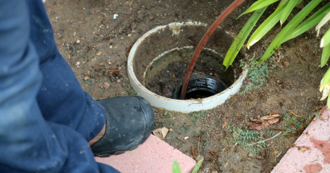How Often Should You Have Your Sewer Lines Inspected? A person in blue pants standing beside an open outdoor drain pipe with a red sewer snake extending inside.