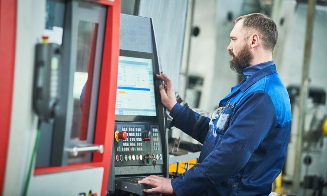 A man stands in front of the control panel and screen of a CNC machine. He looks through various settings.