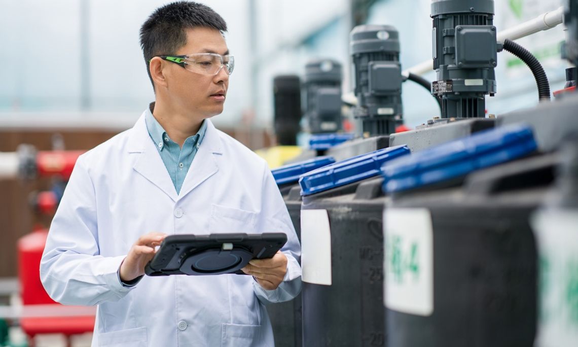 How Protective Coatings Extend Industrial Component Life A man in a long white coat and goggles is observing black and blue containers. He’s holding a tablet in his hand.