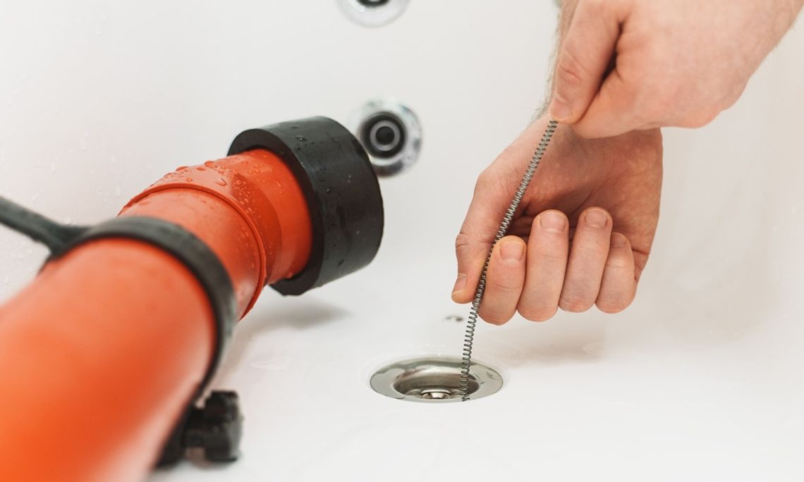 A close-up view shows a person placing a metal drain snake down the drain of a white bathtub. An orange tube is in the tub.