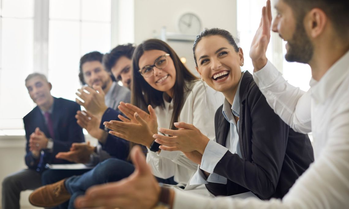 Several people in business attire sitting in a row, clapping and smiling at the person on the end, who has his hands outstretched.