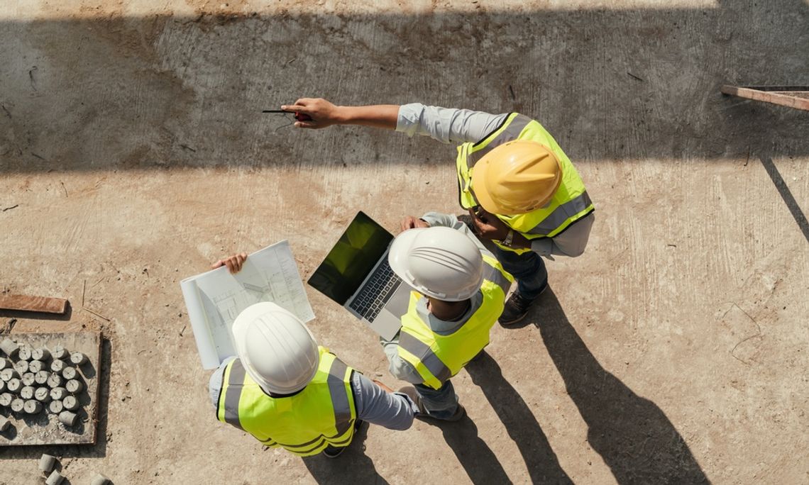 Three construction workers are standing next to each other. One is holding a laptop while another is pointing.