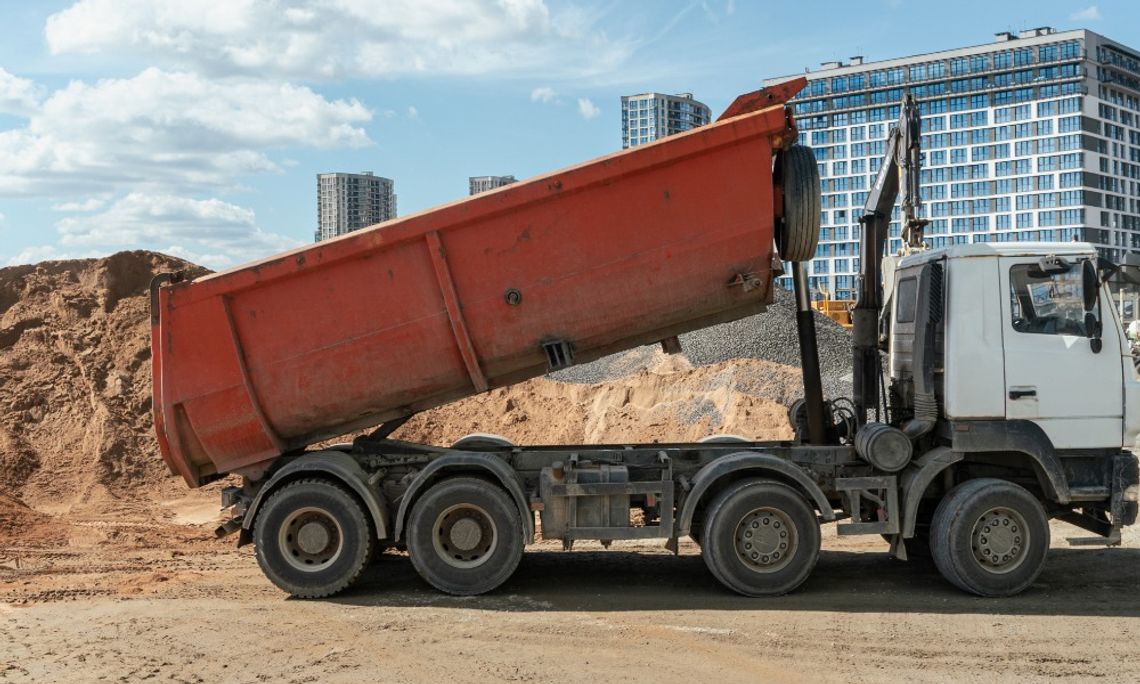 A white dump truck with a raised orange bed standing on dirt beside piles of soil and gravel with city buildings behind it.