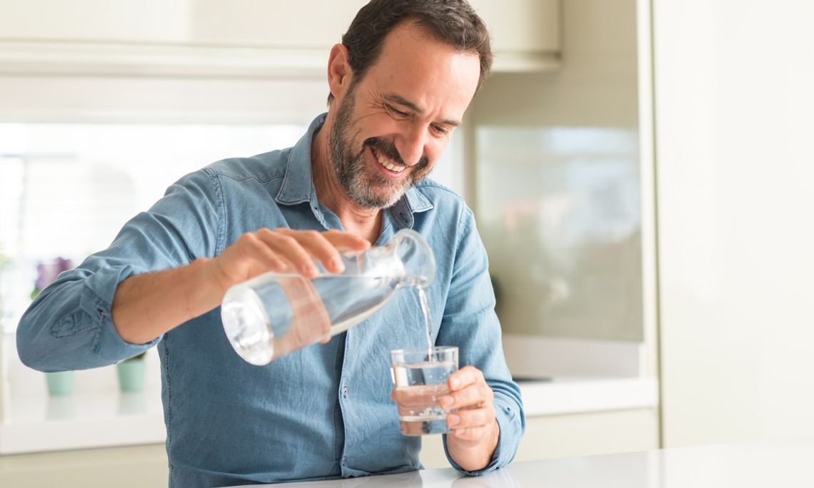 How To Assist Your Body’s Natural Cleansing Processes A man smiles as he stands in his kitchen and pours water from a bottle into a clear glass. He wears a denim shirt.