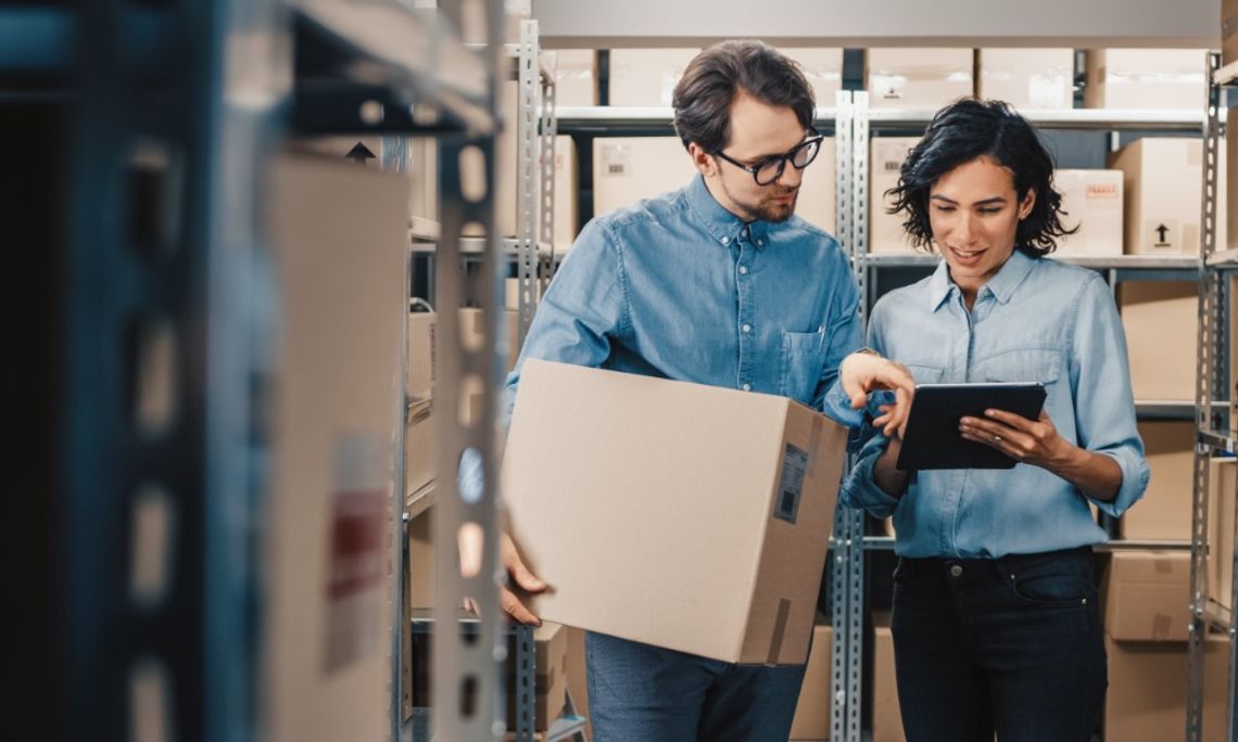 How To Avoid Overstocking Your Warehouse Two employees checking stock levels on a tablet. One employee holds a cardboard box and the other has the tablet.